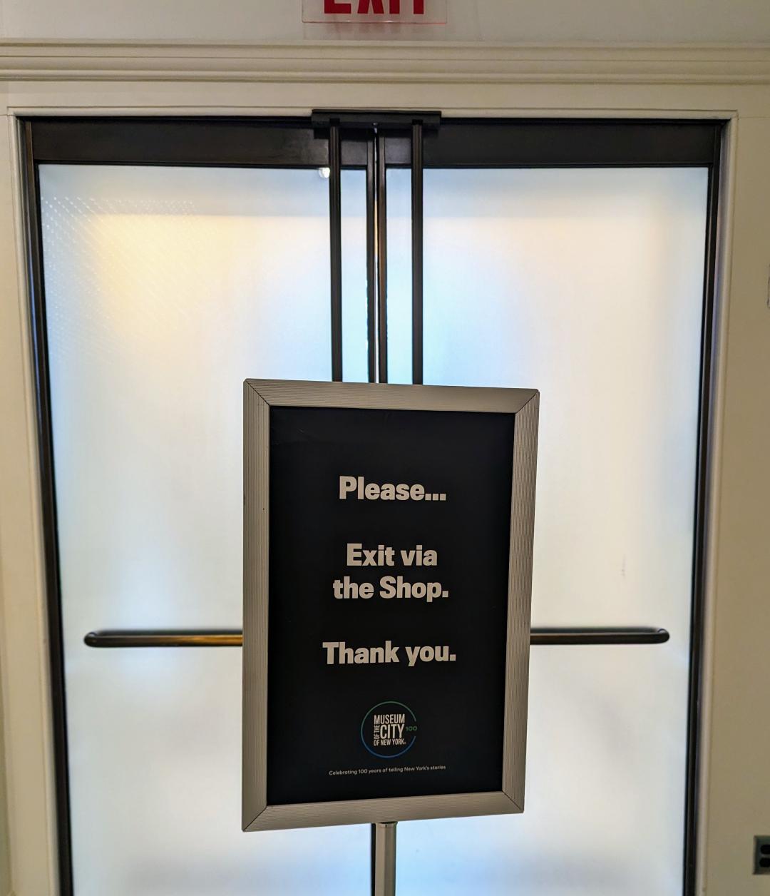 A sign blocking an exit in the New York City Museum, instructing you to exit via the shop