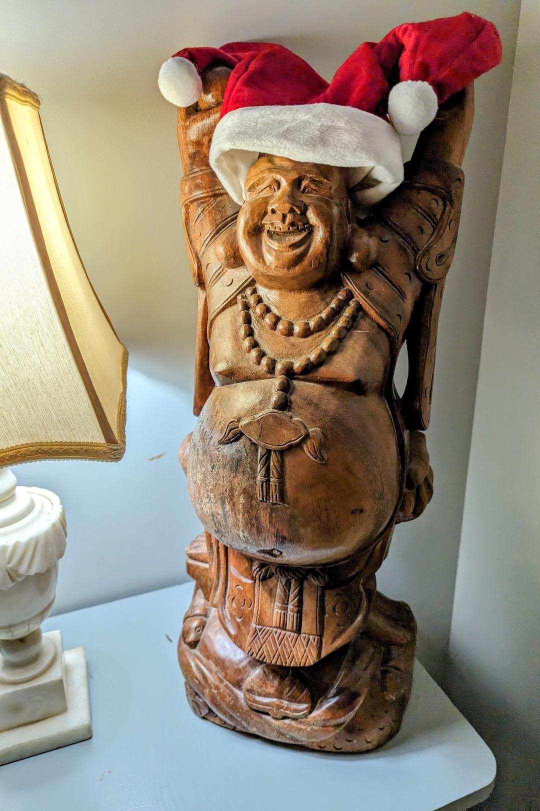 A carved wooden statue of Buddha with a Santa Clause hat placed on Buddha's head