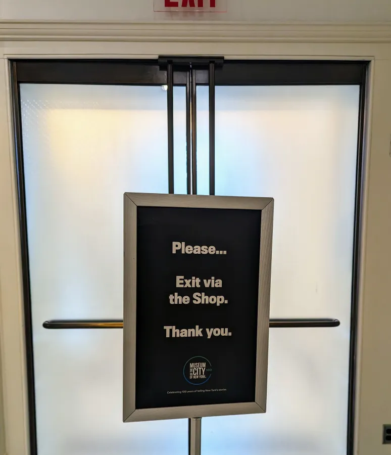 A sign blocking an exit in the New York City Museum, instructing you to exit via the shop