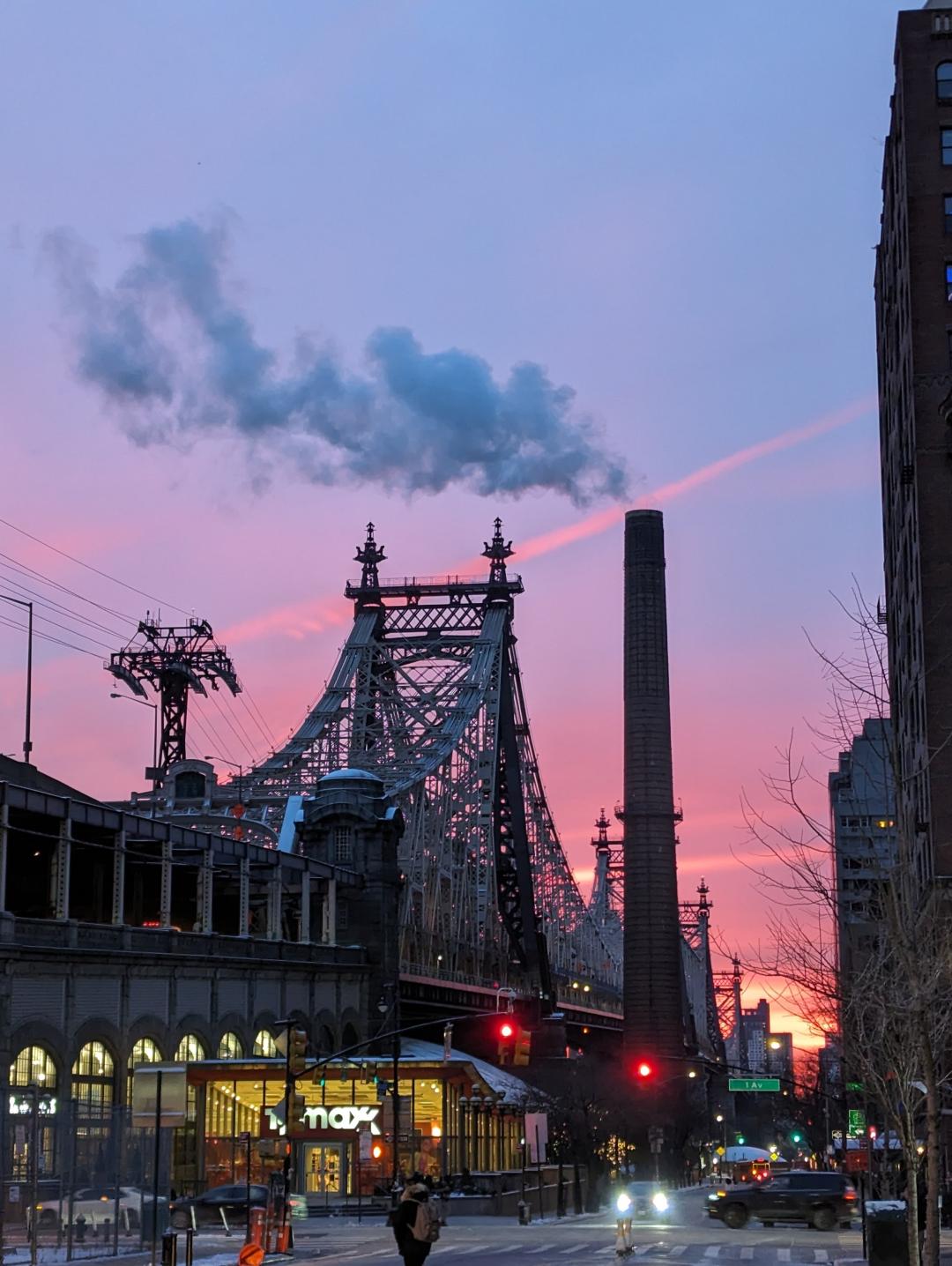 A photograph of the Queensboro Bridge in NYC at Sunrise. The sky is very pink and purple. The bridge is big. A smoke stack is blowing steam. 